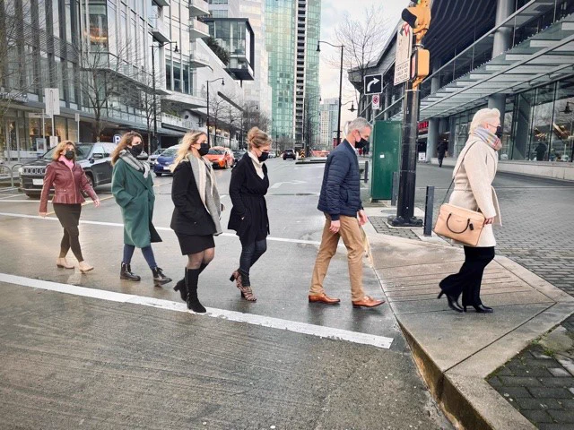 six people in winter outerwear and hygienic face masks crossing the street in a line, reminiscent of The Beatles' Abbey Road album cover.