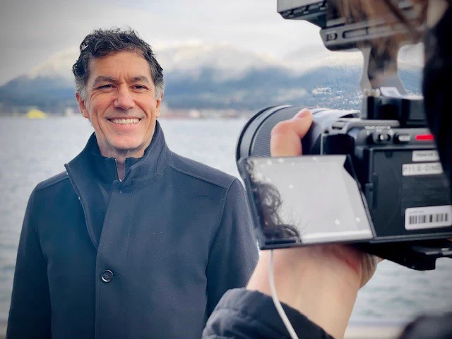 A person with mountains in the far distance behind them looking into a video camera and smiling.