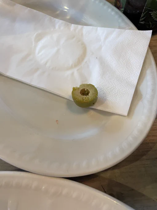 Photo shows a plate with a napkin on it. On the napkin is half a green olive.