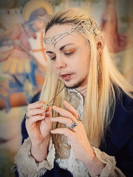 A closer-cropped photo of knightinlace posing in front of a medieval tapestry backdrop, dressed in a blue and silver lord of the rings inspired lolita fashion coord with elf ears and silver circlet, holding up the one ring and hesitating to put it on his finger