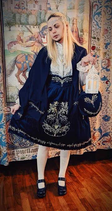 A photo of knightinlace posing with a lantern in front of a medieval tapestry backdrop, dressed in a blue and silver lord of the rings inspired lolita fashion coord with elf ears and silver circlet