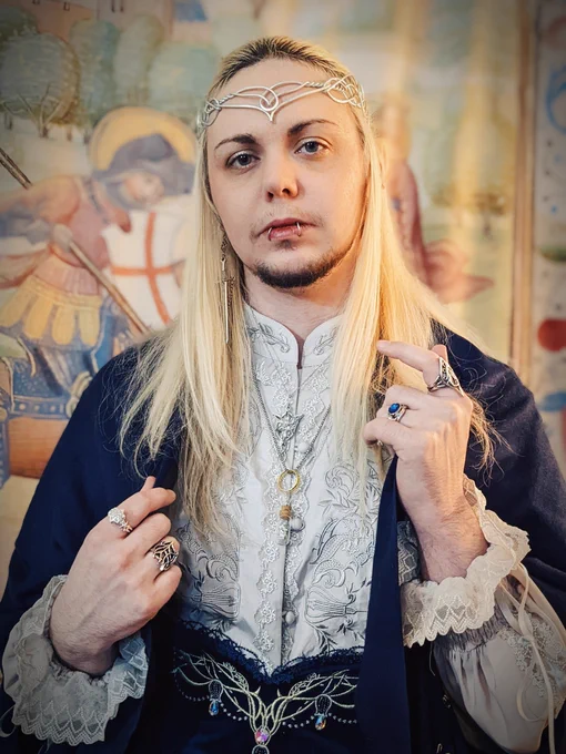 A closer-cropped photo of knightinlace posing in front of a medieval tapestry backdrop, dressed in a blue and silver lord of the rings inspired lolita fashion coord with elf ears and silver circlet