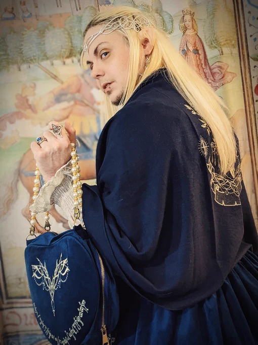 A closer-cropped photo of knightinlace posing in front of a medieval tapestry backdrop, dressed in a blue and silver lord of the rings inspired lolita fashion coord with elf ears and silver circlet, facing away from camera to showcase the embroidered shawl and bag