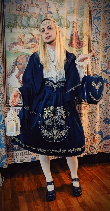 A photo of knightinlace posing with a lantern in front of a medieval tapestry backdrop, dressed in a blue and silver lord of the rings inspired lolita fashion coord with elf ears and silver circlet
