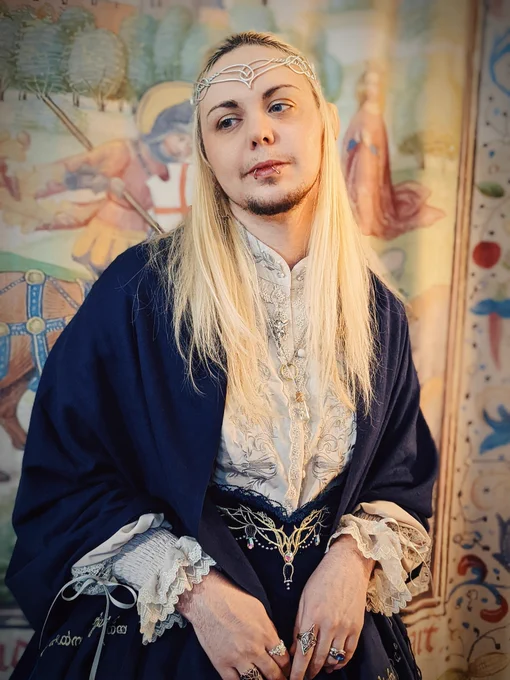 A closer-cropped photo of knightinlace posing in front of a medieval tapestry backdrop, dressed in a blue and silver lord of the rings inspired lolita fashion coord with elf ears and silver circlet