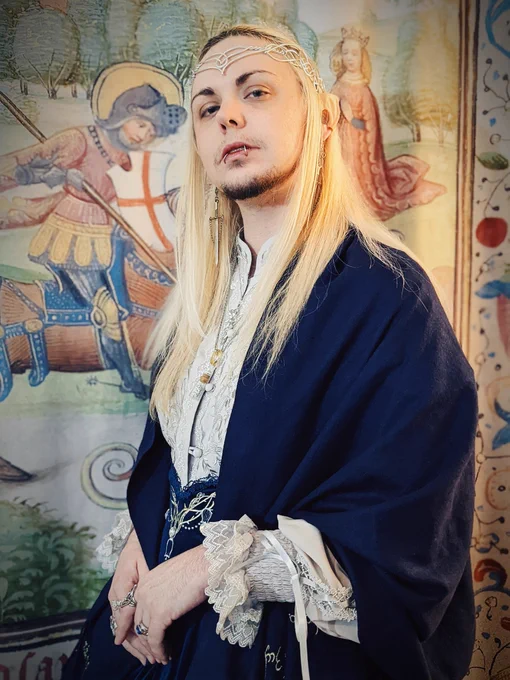 A closer-cropped photo of knightinlace posing in front of a medieval tapestry backdrop, dressed in a blue and silver lord of the rings inspired lolita fashion coord with elf ears and silver circlet