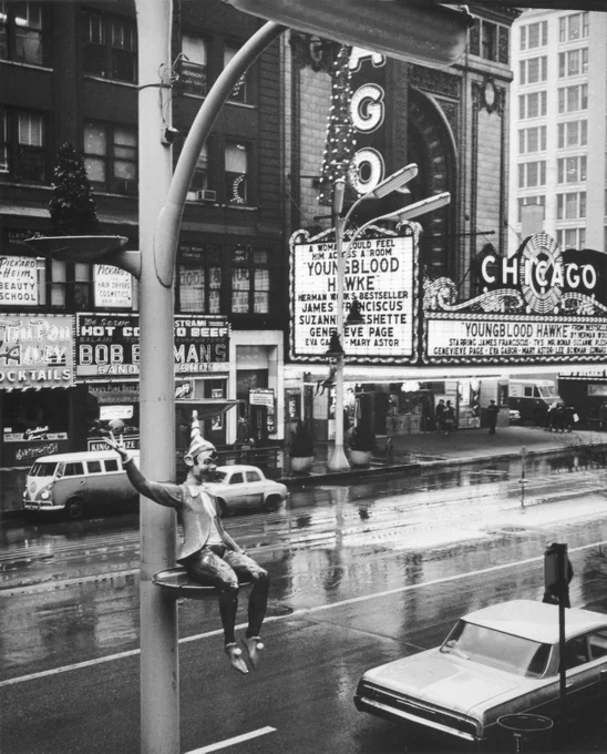 A black & white photograph of an elevated view of a street. there are cards parked on both sides. In the background are tall buildings, which include shops and a marquee and sign for the Chicago Theatre. In the left foreground is a light pole with a small shelf, on which an elf figurine is seated.