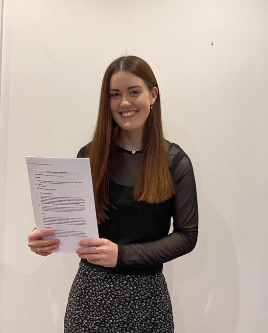 Me standing in front of a wall holding up a piece of paper which is a literary contract. I am wearing a black shirt with white flowers on it and a black top. I also have a silver necklace on. I am smiling and my brown hair is down.