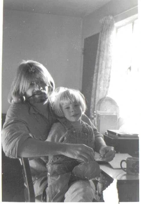 This is a picture of me and my father, circa 1975, in original black & white. I appear about five years old, a white cis boy with straight nearly-white blond hair, bangs cut straight across above my eyebrows. I'm wearing a gray sweatshirt and overalls, probably Oshkosh brand. I'm sitting on my father's lap, one knee forward with foot underneath me, looking down-left toward the camera. My father's arms encircling me appear to be striking a match in front. He and I look much alike, as his only biological son, though his hair is shades darker here and longer, but of a similar shape, as well as nose and mouth and shape of our faces, are all similar. His beard is dark and a little grizzly, and clenched between his teeth is a brown-wood tobacco pipe. His bangs are grown in so the hair has to part above his glasses to see, putting his eyes partially in shadow. He wears a button-up shirt, the sleeves rolled up. Light pours through a window behind us, sitting near his student desk & coffee cup.