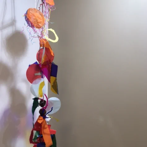 A handmade mobile of coloured acetate shapes, tissue paper, and other materials, hangs from a ceiling casting a shadow on the white wall.  