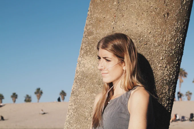 A young woman is leaning against a tree trunk, looking contemplative, on a sunny day with palm trees and sand in the background. She is using a nasal cannula. 