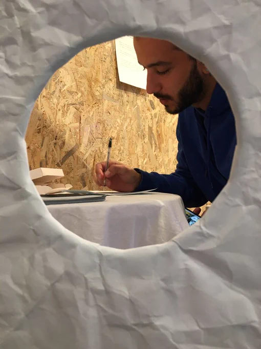 Through a round hole in a wall, a man is seen sat at a desk writing