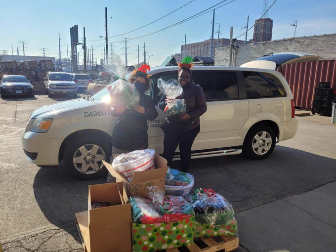A photo of MOPD staff, Sarah and Tyra wearing Elves headbands, standing in from the MOPD van, holding gift bags, standing behind a pallet of gifts.