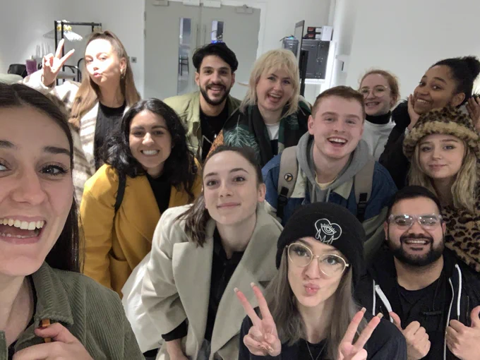 A group of 12 people.  Back row: A woman in a cream & brown coat with brown hair doing a ‘peace’ sign. A man with black hair & a beard in a green jacket, smiling. A person with blonde hair & a fringe in a dark jacket, smiling.  A woman with light brown hair scraped back & glasses wearing a white top, smiling.  A woman with black hair in a high bun, she is smiling with her hand rested on her cheek. Middle row: A woman with black hair down wearing a yellow coat & smiling, a man with short red-ish hair with a blue jacket & a rucksack on, he’s smiling, a woman with blonde hair worn down with a furry leopard print hat & matching jacket, she’s smiling. Front row: a woman with brown hair in a ponytail wearing a cream coat she is smiling, a woman with a black beanie & glasses on with brown hair pouting & doing two peace signs, a man with short black hair and a black beard wearing glasses, smiling with his thumbs up. Front left: a woman with brown hair & a green corduroy jacket, smiling.