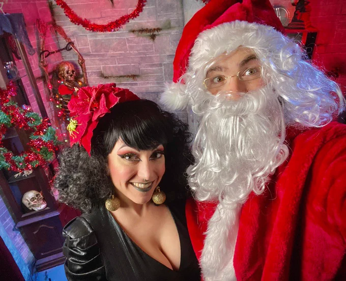 Aurora wearing a low-cut black dress and gold Christmas ornament earrings. she stands next to a Santa Claus with a dungeon scene decorated for Christmas in the background.