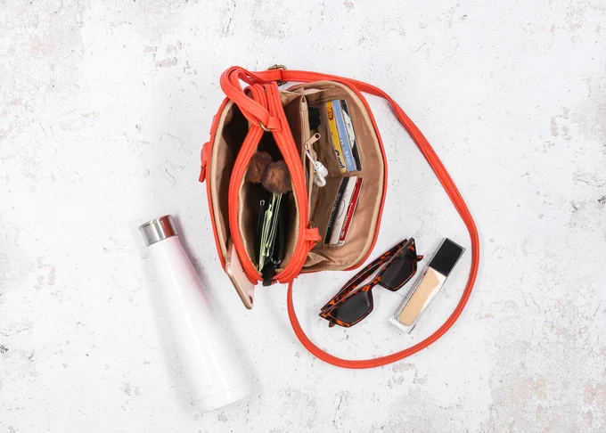 An orange Zippy Triple Compartment Crossbody bag sits open on the countertop showing off the interior contents of the bag which are neatly organized.