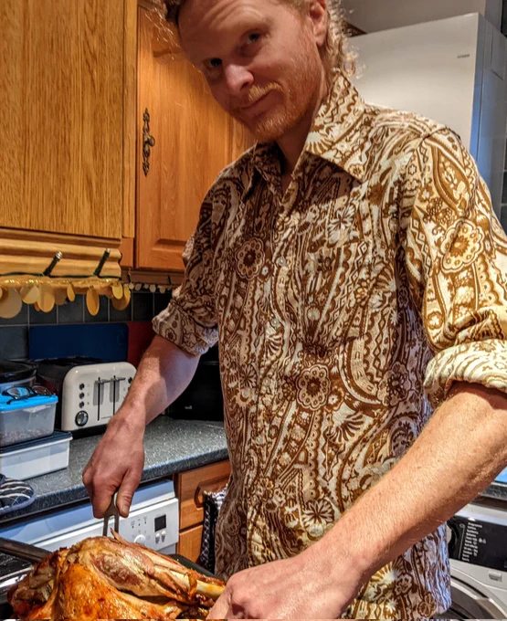 A man wearing a vintage 1970s shirt with a flamboyant brown paisley pattern is carving a turkey