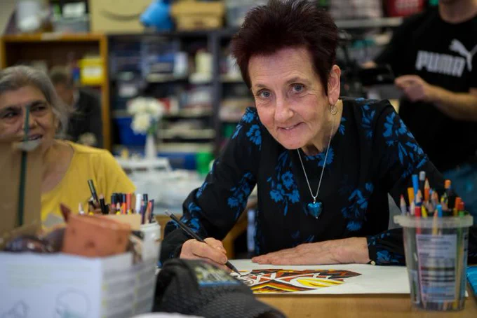 Lucrecia is sitting at a table painting. There are various art materials infront of her