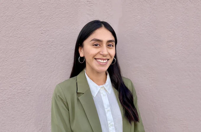 Student in olive green blazer and white button down shirt smiles in front of a blank wall.