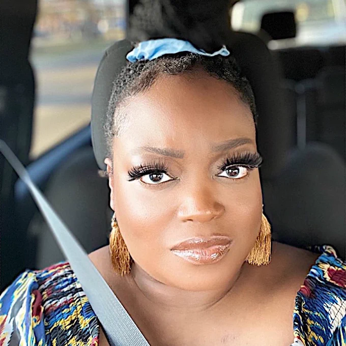 Me, Tinu Abayomi-paul, a dark skinned woman in a vehicle with a grey interior with a grey seatbelt on, a multi-colored scoop neck blouse, a blue scrunchie holding my braided hair atop my head, lookin into the camera with a serious expression. 
