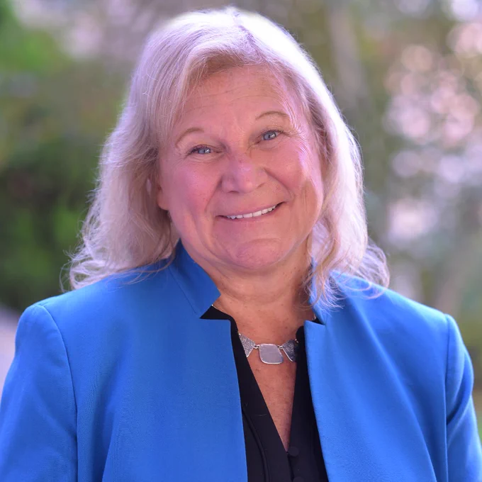 Headshot of CEO Susan Ruzenski: A woman with short blonde hair, blue eyes, black top, blue blazer, and purple necklace.