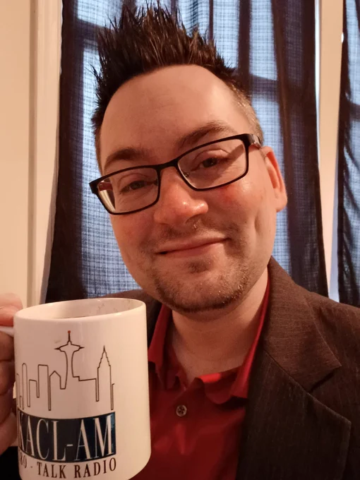Jeff Stormer selfie, in a red dress shirt and grey jacket, holding a KACL-AM Seattle Talk Radio Mug
