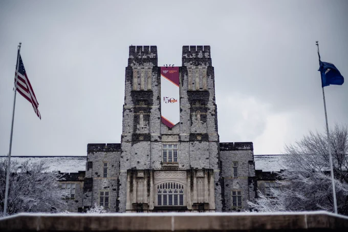 Burruss Hall has two snow covered trees on each side of the building. In front of those trees are flag poles, the pole on the left has the American flag and the pole on the right has the blue Commonwealth of Virginia flag waving in the wind.