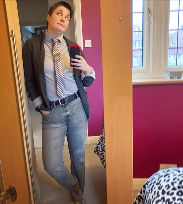 Non in front of a mirror looking up to the left wearing grey jeans, pale grey shirt and striped tie under a khaki cardigan.