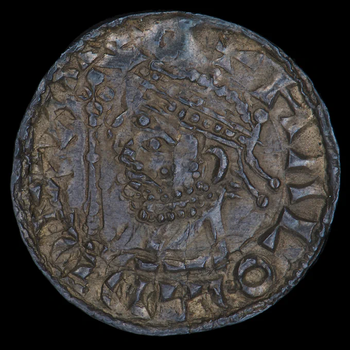 Coin, silver, roughly stamped with a crude profile of a bearded, crowned man. 