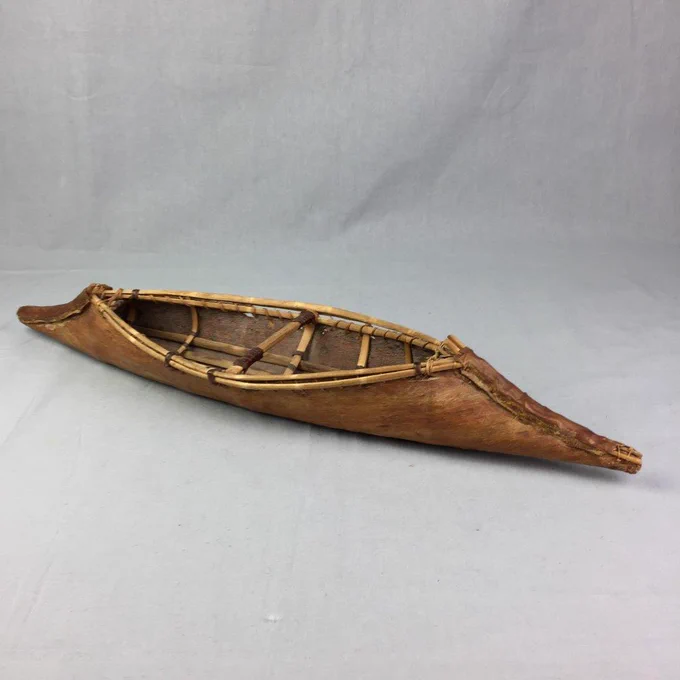 Brown, wooden model of a canoe, photographed on a grey background; the ribs of the canoe are visible on the interior of the model; model is angled on the background