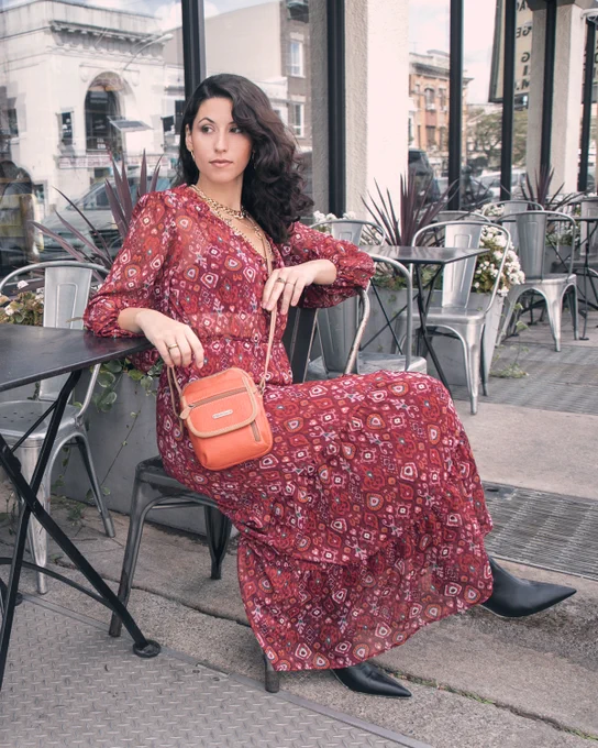 A woman in a red dress sits outside and relaxes waiting for the work day to end. Her orange Micro Everest Crossbody Bag rest by her hip as it hangs from her shoulder.