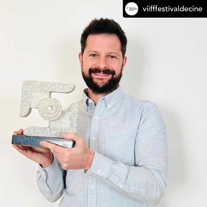 ID: white male with short dark hair and beard wearing a light blue button up shirt holds up a stone award and smiles