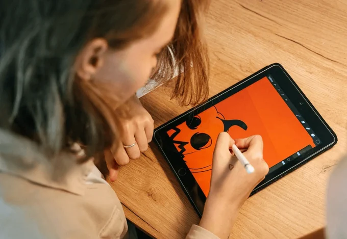 A person with shoulder-length hazel hair is drawing on an ipad. The drawing is of a pumpkin, in black ink on an orange background. The photo is taken over the shoulder of the person. 