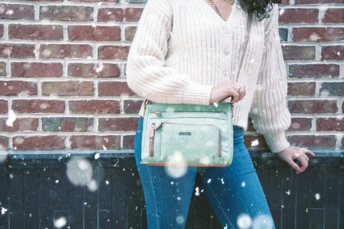 A woman in a white sweater and jeans holds on to her Green Apple East West Summerville Crossbody Bag in today's snow storm.