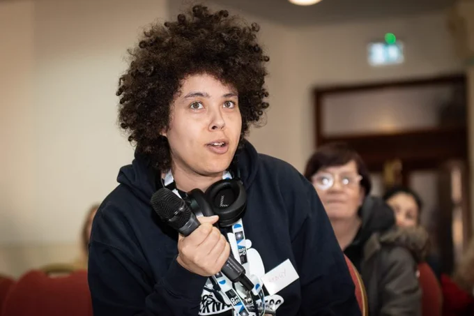Person with dark brown, curly hair wearing a black hoodie holds a microphone slightly away from their mouth, about to speak into it. Or maybe they have just stopped speaking. They have a pair of black headphones and a Coventry City of Culture lanyard around their neck. 