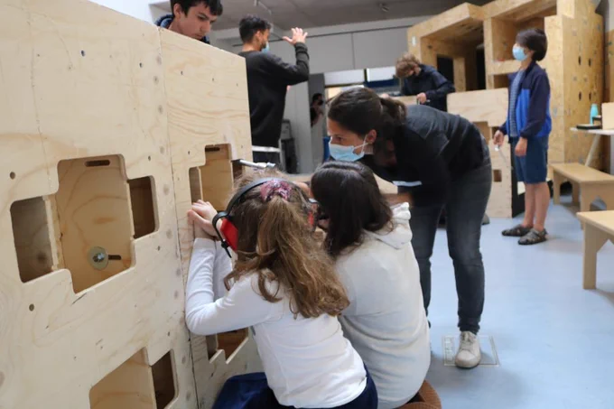 Two young people with long brown hair and white t-shirts are seem to be screwing something into a wooden pallet, supported by an older person wearing dark jeans and a tshirt, wearing a surgical facemask. There are other people in the background doing similar work. 