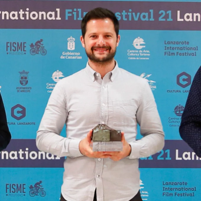 A man smiles holding an award, behind him a blue backdrop for the Lanzarote International Film Festival​