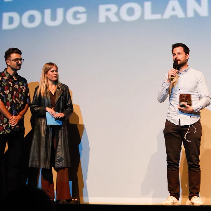 Same man on stage speaking into a microphone. To his right, a man and a woman stand watching.​