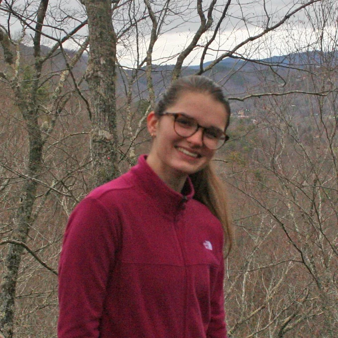 Brannan standing in front of a forest in the winter (no leaves on trees) with mountains in the background