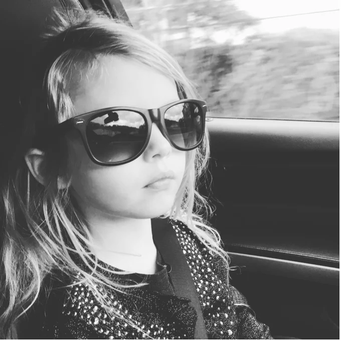 A black and white photo of a beautiful young girl with long blonde hair sitting in a car. She is wearing dark sunglasses and a spotty top, she has a seatbelt over her shoulder. You can see trees and a hedge out of the window behind her.