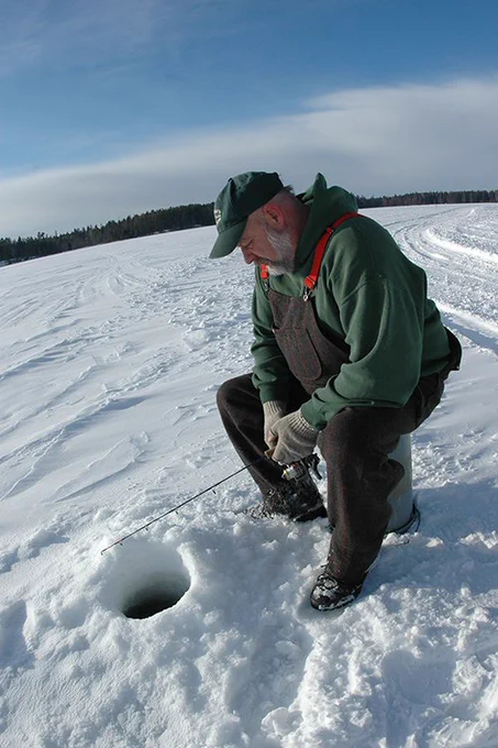 A man sitting on an overturned bucket on the ice. He is holding a fishing pole over a hole in the ice.