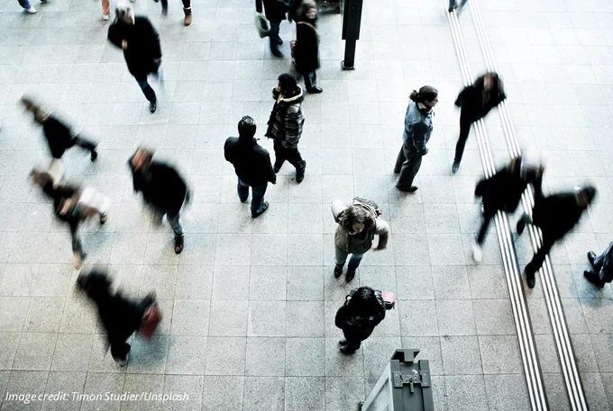 Photograph of a crowd of people in a public area or square, walking in different directions, some of them blurred by motion. Image credit: Timon Studier/Unsplash