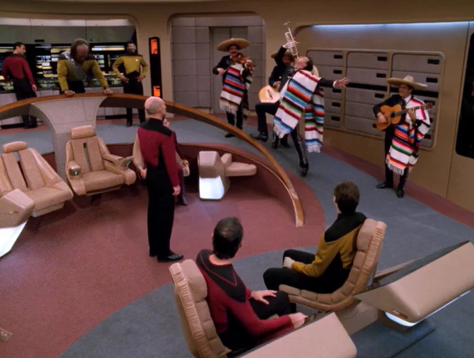 Screenshot of Star Trek The Next Generation. Interior, Enterprise Bridge. The bridge crew members are looking at Q who is playing a trumpet while wearing a striped Mexican scarf. The colors of Q's scarf are red, blue, green, black, and white, so they match the scarves of the rest of the mariachi band. Three mariachi band members are playing guitars and a violin behind Q. Picard is staring at Q from center stage. La Forge is looking at Q from a rear science station, not wearing any hat. Worf is at his post, looking down, frustrated, his hands resting on the railing. Data is at navigation, turned around in his chair to look at Q. An ensign is at the conn, turned in his chair to look at Q. Small black spots on the wall behind Q are missing their little lights. A tall lighted alert display behind La Forge has its lights at the top and bottom instead of near the center. Riker is missing from the picture.