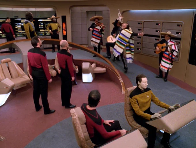 Screenshot of Star Trek The Next Generation. Interior, Enterprise Bridge. The bridge crew members are looking at Q who is playing a trumpet while wearing a striped Mexican scarf. The colors of Q's scarf are ochre, purple, blue, black, and white, so they do not match the scarves of the rest of the mariachi band. Three mariachi band members are playing guitars and a violin behind Q. Picard is staring at Q from center stage. La Forge is looking at Q from a rear science station, wearing a sombrero like the mariachi band members. Worf is at his post, looking behind himself at La Forge. Data is at navigation, facing towards his console, ignoring Q. An ensign is at the conn, turned in his chair to look at Q. Small black spots on the wall behind Q are lit with little lights. A tall lighted alert display behind La Forge has its lights near the center, with blank areas at the top and bottom. Riker is standing behind Picard, looking at Q.