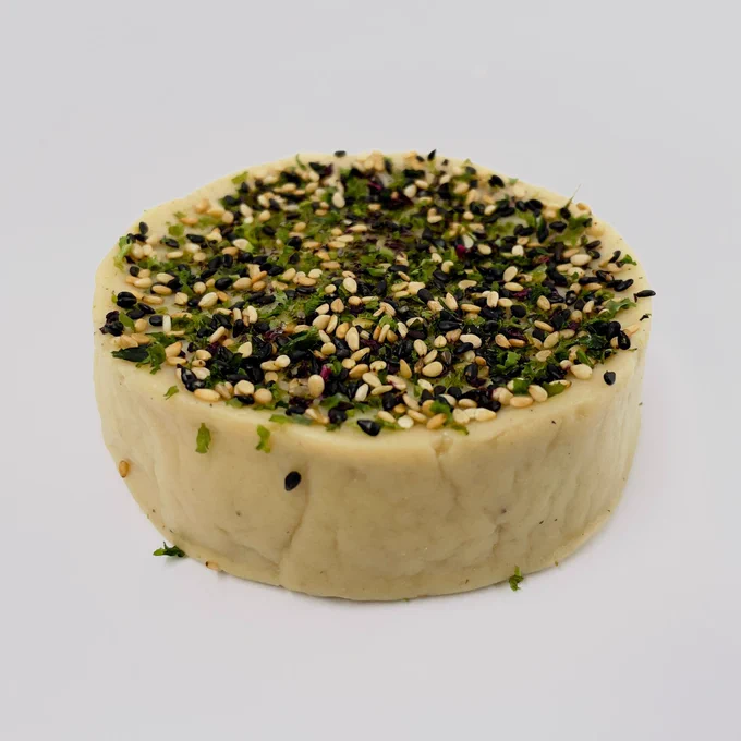 A truckle of vegan cheese topped with black and white sesame seeds and flakes of seaweed.