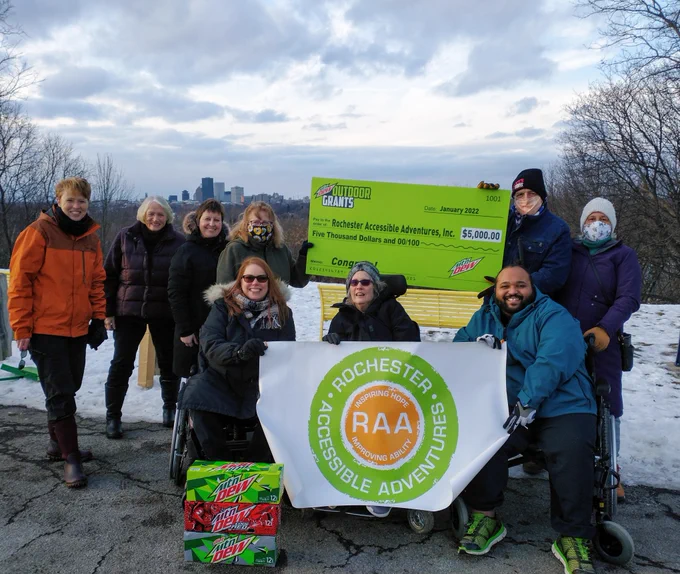 Group holding a large bright green Mtn Dew Check and an RAA logo banner, standing at Cobbs Hill Reservoir overlooking the City of Rochester, six people standing, three people in wheelchairs -- all smiling!