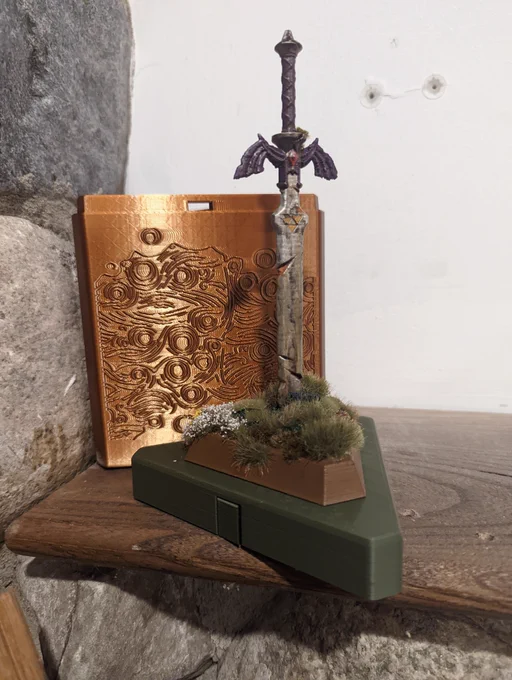 Photo of a 3D Printed Zelda themed Twist Lock Box. The box has a painted and weathered Master Sword topper stuck into the ground with fake bushes and flowers around it.