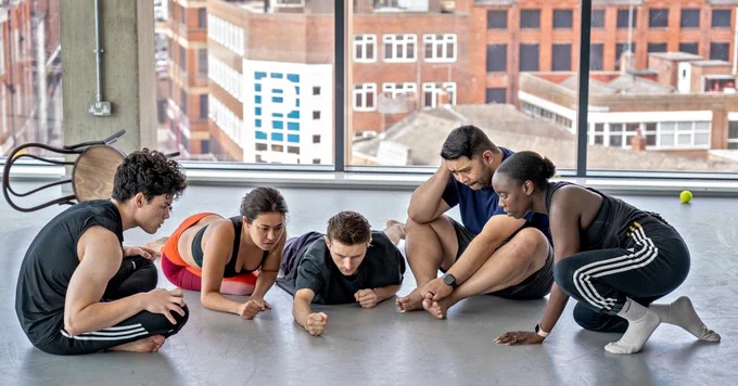 Five performers sitting low on the floor in a semi-circle in a bright rehearsal room with big windows overlooking Leeds city centre.