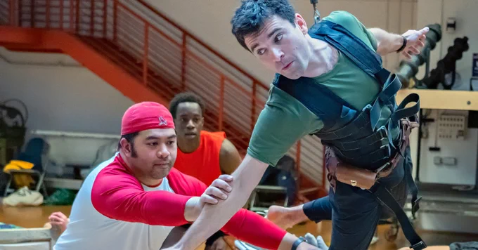An actor playing Peter Pan flying in a harness in the rehearsal room with other performers helping to keep him steady.