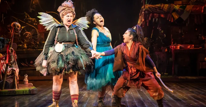 Actors on stage in costumer as Tinkerbell, Wendy and Tiger Lily. They are touching hands, have their mouths open and have strong expressions on their faces as if swearing allegiance to one another.